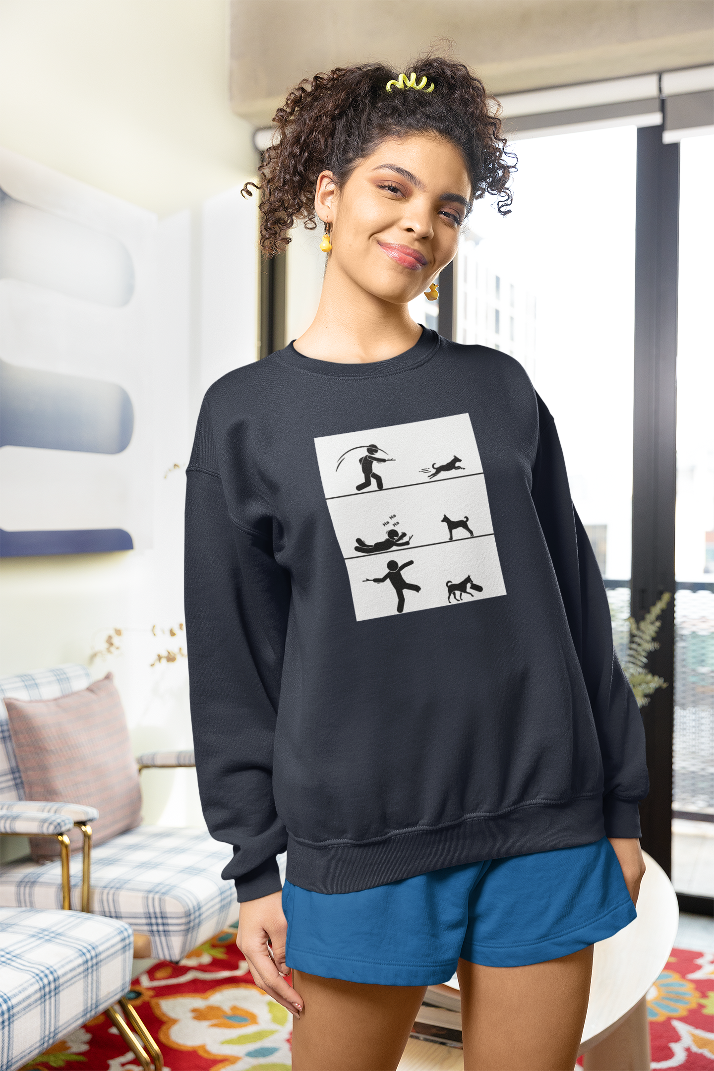 A woman is wearing a black crewneck sweatshirt with a graphic design in the center, smiling and posing in a room with light decor.