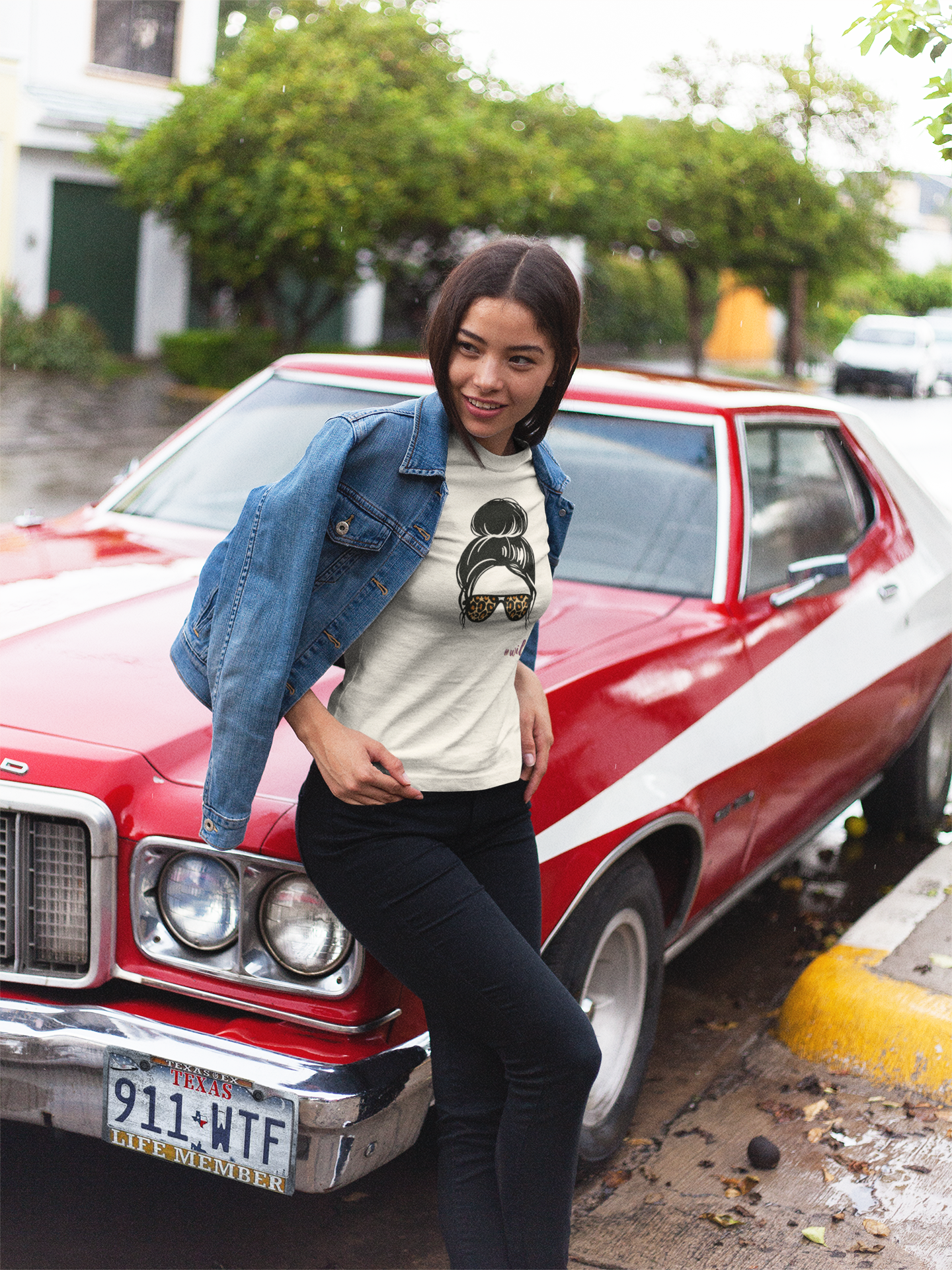 Woman standing next to a red car with a white stripe, wearing a graphic t-shirt and denim jacket.